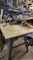 Full view of radial arm saw mounted on wooden work table with metal legs, showing saw blade and arm controls.