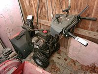 Front-side view showing engine housing, fuel tank with red cap, wheels and auger cover