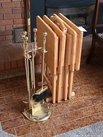 Photo showing four wooden folding TV trays stored in a wooden stand beside a brass fireplace tool set on a tiled floor near a fireplace.