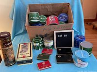 Full assortment of assorted vintage and new tins in and around a cardboard box on blue drape background