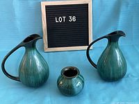 Three vases arranged in front of a blue cloth background with a small board marked 'LOT 36'. Two tall vases with handles and one small round vase, all green in color with glaze showing gradient shades.