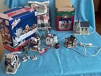 Image showing all porcelain Christmas village houses, accessory figurines, miniature trees, and original packaging from Zellers and Santa's Trim Shoppe. Also visible is a bag with Christmas tablecloths and runners.