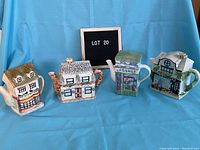 Four ceramic collectible teapots shaped like village buildings displayed side by side against a blue backdrop with a sign labeled 'LOT 20'.