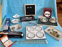 Wide shot showing assortment of entertaining and decorative items on blue cloth backdrop including crystal coasters, porcelain cups and saucers, decorative bowls, tongs and ornamental glass item.