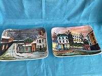 Two rectangular decorative plates painted with cityscapes, shown side by side on a blue cloth background.