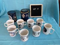 Front view of eight assorted Charles and Diana royal collectible mugs and two tins on blue fabric background with a letter board displaying 'Lot 17'.