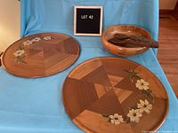 Photo displaying two large round wooden trays with geometric segmented wood patterns and floral painted designs, alongside large wooden salad bowl and tongs.