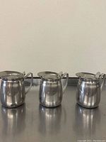 Front view of 3 stainless steel milk frothing pitchers showing handles and hinged lids closed.