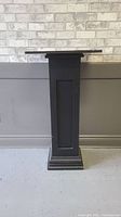 Front view of the black wooden decorative column stand showing rectangular top, paneled inset sides, and stepped base.