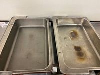 Two full size stainless steel food pans side by side on table, showing front and top view, with visible wear and stains.