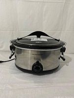 Front and side views of Hamilton Beach Stay or Go 6-quart slow cooker showing stainless steel body, control dial, glass lid with clip-tight seal, and full-grip handles.