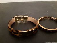 Photo showing wide cuff-style leopard print bangle and slimmer textured gold-tone bangle resting on black surface with Fossil stamp visible on clasp of larger bangle.