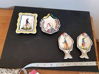 Four small decorative Quimper plates arranged on black background with measuring tape for scale. Two fish-shaped plates and two scalloped edge plates with hand-painted Breton folk art figures.
