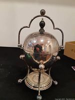 Front view of the antique silver egg coddler closed with lid on stand with burner base.