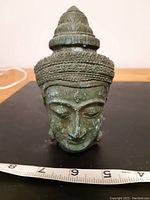 Front view of the antique metal Buddha head showing detailed facial features and headpiece with measuring tape indicating approximately 7 cm in height.