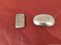 Two antique items on red textured surface; match stick holder engraved with floral scrollwork and plain oval snuff box