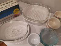 Photo showing two clear glass baking dishes, one with lid, square dish with ribbed base, white ramekin, small ceramic floral bowls, and blue glass bowls, with food sealer box in background.