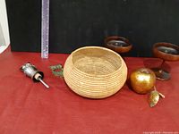 Photo showing the First Nations basket center, wooden wine cups back, metal apple, and metal spout on red surface with ruler for size reference.
