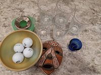 Wide top view showing all items together on marble surface: clear glass tumblers, yellow pottery bowl with golf balls, African mask, and green glass hair receiver.