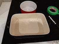 Rectangular beige Maxwell Williams casserole dish with brown handles and round red casserole dish behind it.