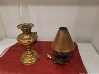 Overview of both lamps including brass oil lamp with glass chimney and copper electric lamp with conical shade on fabric surface.