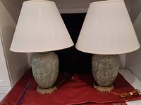 Pair of celadon pottery lamps with cream lampshades on red fabric background.