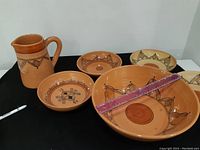 Photo showing six pottery items including one jug and five bowls with various black painted geometric and floral designs on terracotta clay.