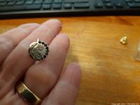 Front view of small round 10K gold tie pin held between fingers showing fish design and jewel border.