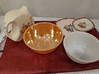 Photo showing all items on a red cloth: the Universal Cambridge pitcher with floral pattern, orange and white Fire King bowls, and two floral square plates.