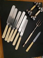 Photo of six butter knives, one smaller knife, two utensil rests and the fork laid out on a dark surface.