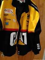 Front side of black, yellow, and red racing jacket with CAT logo and multiple NASCAR-related patches.
