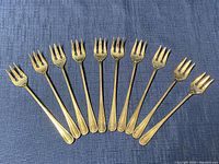Full view of ten vintage cocktail forks arranged in a row on a blue textured fabric background, showing consistent design and condition.
