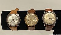 Three men's automatic watches displayed on black cushion. Includes two Rona watches and one Timex watch with leather straps in brown and tan, silver and gold dials showing age patina and wear.