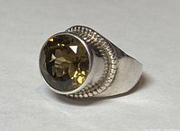 Close-up photo of silver ring head showing large round faceted yellow-brown colored crystal set in bezel with surrounding textured border.