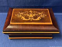 Side angle view of the wooden jewelry box showing the decorative urn and floral design on the lid and the curved molding around the edges.