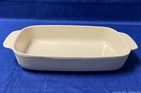 Side view of cream-colored rectangular cast iron enamel baking dish on blue fabric background showing integrated handles and smooth interior.