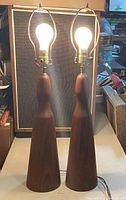 Pair of teak wood table lamps with light bulbs attached, showing front profile with natural wood grain and brass harp fittings.