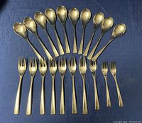 Top-down view of flatware set laid out on blue cloth background. Includes 7 dinner forks, 1 salad fork, 2 luncheon forks in front row, 5 dinner spoons and 5 teaspoons in back row. Pieces have a brushed matte finish with minor wear.