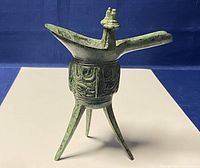 Front angle view of bronze ritual wine vessel reproduction highlighting the spout, tripod legs, decorative body, and finials.