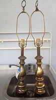 Pair of tall brass table lamps shown standing inside a metal sink. Each lamp has a rounded tiered base, a brass harp, and a finial on top. No shades are present.