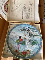 Photo of a Legends of West Lake series decorative plate featuring two women in traditional Chinese dress near a scenic outdoor setting with flowers and trees, with certificate paperwork visible inside the original box.