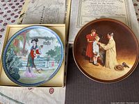 Two collectible plates displayed with certificate and boxes. One plate depicts traditional Chinese woman in garden scene (Beauties of the Red Mansion). The other plate depicts a man and woman with a child in a diagnostic setting titled 'Ready for the World' by Norman Rockwell.