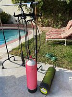 Two diving tanks standing outdoors near a swimming pool. One has red mesh cover and regulator attached, the other is black mesh-covered lying horizontally.