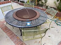 View of the full outdoor wrought iron patio set with round fire pit table and 4 matching chairs on a concrete patio.