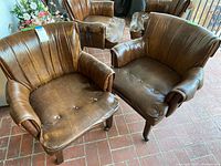 Pair of brown vintage vinyl club chairs with worn upholstery and button tufting, shown on brick floor with some wear and tear visible.