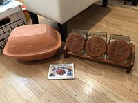 Photo showing terracotta Schlemmer Topf roaster with embossed designs and three vintage glass canisters with wood lids on a wooden rack, also Schlemmer recipe booklet visible