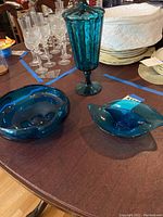 Three pieces of aquamarine teal blue glassware on wooden table: two curved bowls and one tall covered pedestal jar.