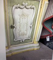 Close-up of ornate carved panel on cabinet door showing distressed paint in cream and green with yellow accents.