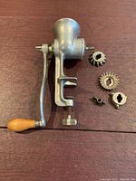 Photo of vintage meat grinder body with attached wooden handle and three separate grinding plates and a wing-shaped attachment pieces on table.