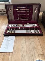 Full view of the silver-plated flatware set inside a wooden presentation box with a plush maroon velvet lining. Pieces include forks, spoons, knives, and serving utensils secured in their slots.